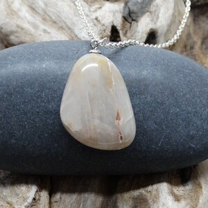 May include: A silver chain necklace with a light brown, smooth, oval-shaped pendant. The pendant has a subtle, white, marbled pattern.
