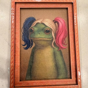 May include: Framed artwork featuring a green frog with blonde and pigtails, one blue and one pink. The frog is wearing a costume. The frame is orange with a textured pattern. The artwork is in a clear plastic sleeve.