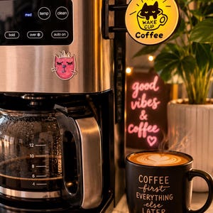 May include: A stainless steel coffee maker with a digital control panel, a glass carafe, and a circular yellow sign reading WAKE CUP Coffee. A black mug with the text COFFEE first EVERYTHING else LATER and a heart icon sits next to the machine.