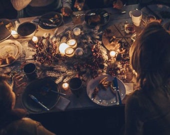 Op de afbeelding: Een feestelijke dinertafel met borden, bestek en brandende kaarsen. De tafel is versierd met groen en bessen, wat een warme sfeer creëert. Mensen verzamelen zich rond de tafel en genieten van een maaltijd.