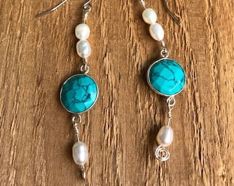 Op de afbeelding: Een paar turquoise en witte hangende oorbellen. De oorbellen hebben een turquoise steen met een zilveren zetting en een witte parel.