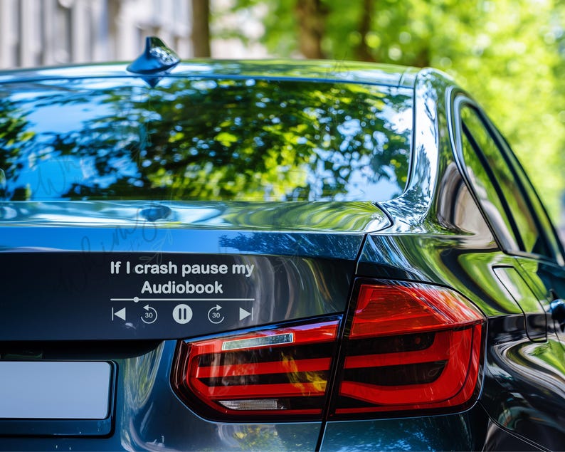 May include: A black car's rear with a white decal that reads "If I crash pause my Audiobook" above a media player interface. The car's taillight is red and the background is blurred green.
