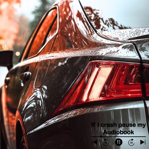 May include: Close-up of a dark gray car's rear, with a red taillight and the text "If I crash pause my Audiobook" with a media player graphic. The car is wet, reflecting the surrounding autumn foliage.