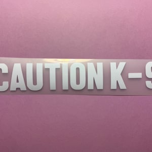 May include: A rectangular sticker with the words "CAUTION K-9" in bold, white capital letters. The sticker is on a light pink background. The text is centered and clearly visible.