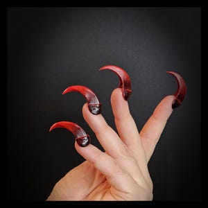 May include: Four red and black claw-like fingernail tips, worn on a hand, with the tips curved and pointed.