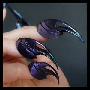 May include: Close-up of a hand with three long, black and green, claw-like nail extensions.