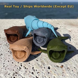May include: Four articulated whale shark toys in light blue, dark gray, brown, and olive green. Each figure features a wide, open mouth with visible teeth and a textured, scale-like surface. The text Real Toy / Ships Worldwide (Except EU) is visible.