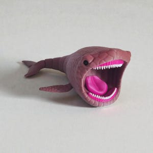 Puede incluir: Un juguete caprichoso en forma de ballena de color malva, con la boca muy abierta que revela encías y lengua de color rosa brillante, y dientes blancos. La ballena tiene aletas pequeñas y una cola, y una piel texturizada. El juguete está sobre un fondo liso y claro.