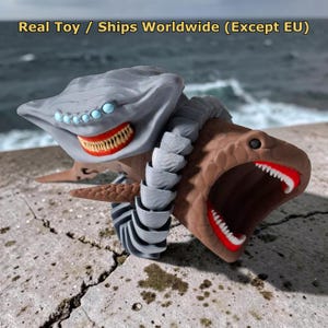 May include: A gray and brown articulated toy featuring two connected creature heads with multiple light blue eyes, sharp white teeth, and red mouths. The text "Real Toy / Ships Worldwide (Except EU)" is visible at the top of the image.