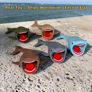 May include: Five shark toys in olive green, tan, brown, gray, and light blue. Each shark features an open mouth with white teeth and a bright red tongue. The text "Real Toy / Ships Worldwide (Except EU)" is visible at the top of the image.