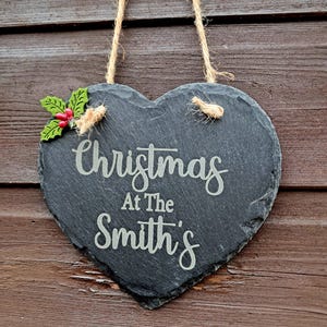 May include: Heart-shaped slate sign with the text "Christmas At The Smith's" in white script. The sign is decorated with a sprig of holly and hangs from a jute rope. The sign is hanging on a wooden background.