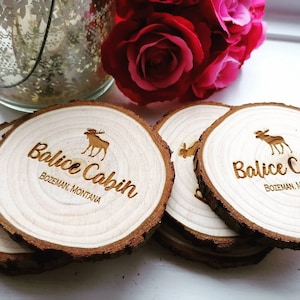 Könnte beinhalten: Vier Untersetzer aus Holz mit rustikalem, natürlichem Finish. Jeder Untersetzer ist mit einem lasergravierten Bild eines Elches und dem Text "Balice Cabin, Bozeman, Montana" versehen.