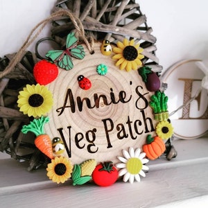 Pode incluir: Uma placa de madeira com o texto "Annie's Veg Patch" em letras pretas. A placa é decorada com flores, vegetais e insetos de feltro coloridos.
