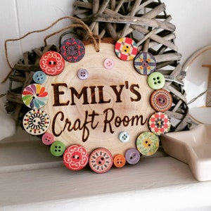 Könnte beinhalten: Ein Holzschild mit dem Text "Emily's Craft Room" eingebrannt in die Oberfläche. Das Schild ist mit bunten gemusterten Knöpfen am Rand verziert.