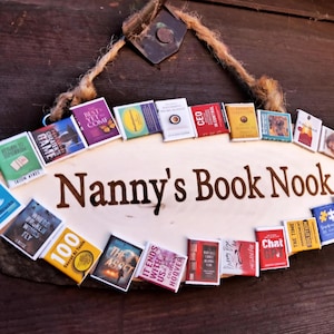 May include: A wooden sign with the text "Nanny's Book Nook" surrounded by a variety of colorful book covers. The sign is hanging on a wooden wall.