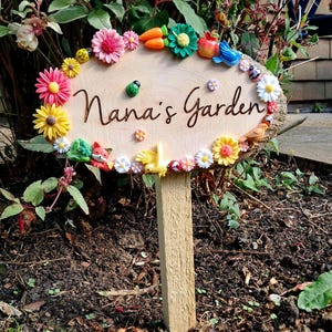 Puede incluir: Un letrero de jardín de madera con el texto "Nana's Garden" rodeado de coloridas decoraciones de flores y animales. El letrero está montado en una estaca de madera y colocado en un jardín.