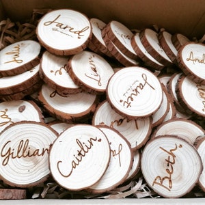 Puede incluir: Una colección de rodajas de madera redonda con nombres escritos en letras doradas. Los nombres incluyen Jared, Gillian, Caitlin, Beth y Liam. Las rodajas están dispuestas en una pila, con algunas superpuestas.
