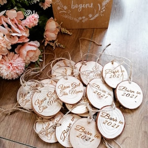 May include: A collection of wooden ornaments with various sayings, including "You Got This Mama" and "Born in 2021". The ornaments are made of natural wood and have a rustic look.