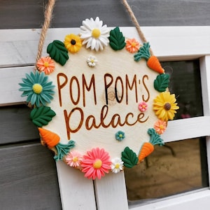 May include: A wooden sign with the words "Pom Pom's Palace" written in black. The sign is decorated with colourful flowers and carrots.