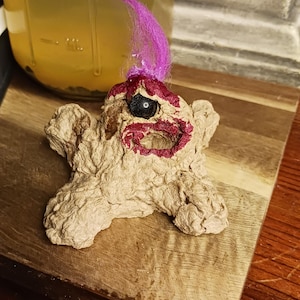 May include: A handmade, textured papercraft figure with a cross-like shape. It features a painted face with a black eye, red lips, and purple hair. The figure rests on a wooden surface.