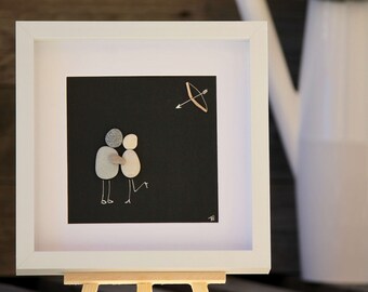 以下が含まれることがあります： 黒い背景に、小石で描かれた抱き合う2人の姿が特徴の額装されたアートワーク。その上に、木の弓矢が描かれています。アートワークは白いフレームに入っており、小さな木製のイーゼルに飾られています。