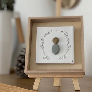 May include: A framed artwork featuring a hand-painted stone figure with a brown face and a gray body. The figure is surrounded by a black and white floral wreath. The artwork is displayed on a wooden easel.