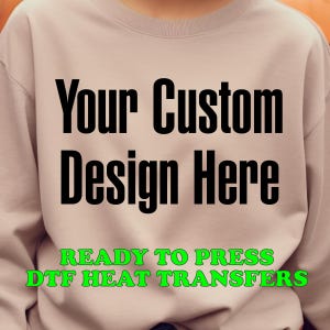 May include: A light pink sweatshirt with the text "Your Custom Design Here" in black and "READY TO PRESS DTF HEAT TRANSFERS" in green.