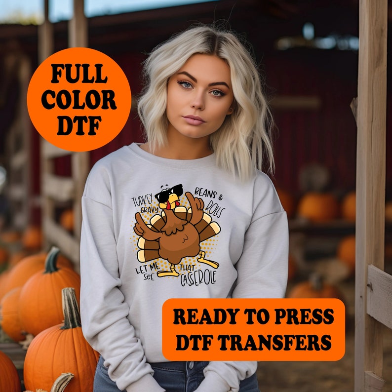 Op de afbeelding: Een grijs sweatshirt met een Thanksgiving-thema ontwerp met een cartoon kalkoen met een zonnebril en de tekst "Turkey Gravy", "Beans & Rolls", "Let Me See" en "That Casserole". De tekst "Ready To Press DTF Transfers" is in oranje op een witte achtergrond gedrukt.