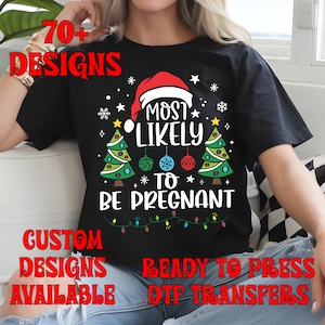 May include: Black t-shirt with the text "MOST LIKELY TO BE PREGNANT" in white, with a Santa hat, Christmas trees, and ornaments. The shirt also has the text "70+ DESIGNS", "CUSTOM DESIGNS AVAILABLE", and "READY TO PRESS DTF TRANSFERS".