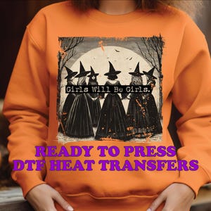 May include: Orange sweatshirt with a graphic of five witches in black dresses and pointy hats. The graphic has a distressed look and the text "Girls Will Be Girls."  The sweatshirt also has the text "Ready to Press DTF Heat Transfers" in purple.