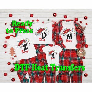 May include: Four white t-shirts with red and white plaid pants. The shirts have a Christmas light design with a Santa hat and a letter on each shirt. The shirts say "Dad", "Mom", "Zoey", and "Lexi". The text "Ready to Press DTF Heat Transfers" is at the bottom of the image.