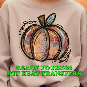 May include: A light brown sweatshirt with a tie-dye pumpkin design and gold glitter accents. The text "READY TO PRESS DTF HEAT TRANSFERS" is printed in green below the pumpkin.