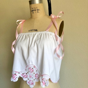 May include: White crochet top with pink floral details and pink satin ribbon straps. The top has a scalloped hem and is on a dress form.