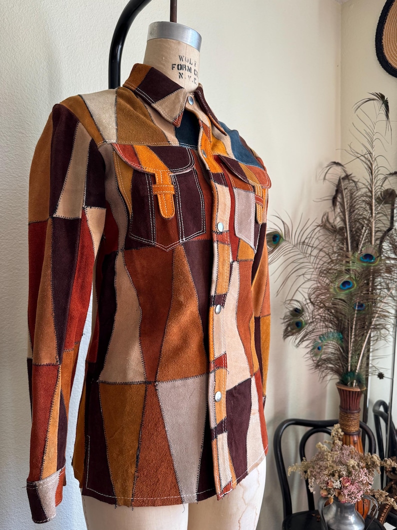 Pode incluir: Uma camisa de camur&ccedil;a em patchwork com fecho de bot&otilde;es e mangas compridas. A camisa &eacute; feita de diferentes pe&ccedil;as de camur&ccedil;a de cores diferentes, incluindo castanho, bege e laranja. A camisa tem um colarinho e um bolso no peito esquerdo.