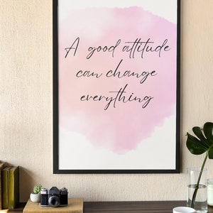 Peut inclure: Une impression encadr&eacute;e avec un fond aquarelle rose et le texte "A good attitude can change everything". L'impression est sur un bureau en bois avec un ordinateur portable, un appareil photo, une plante en pot, une tasse et une soucoupe, et un vase avec une plante.