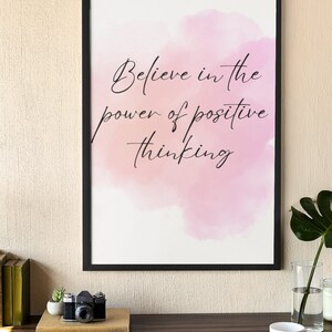 Peut inclure: Une impression encadr&eacute;e avec un fond aquarelle rose et le texte "Croyez au pouvoir de la pens&eacute;e positive". L'impression est sur un bureau avec un ordinateur portable, un appareil photo, une plante en pot et une tasse et une soucoupe.