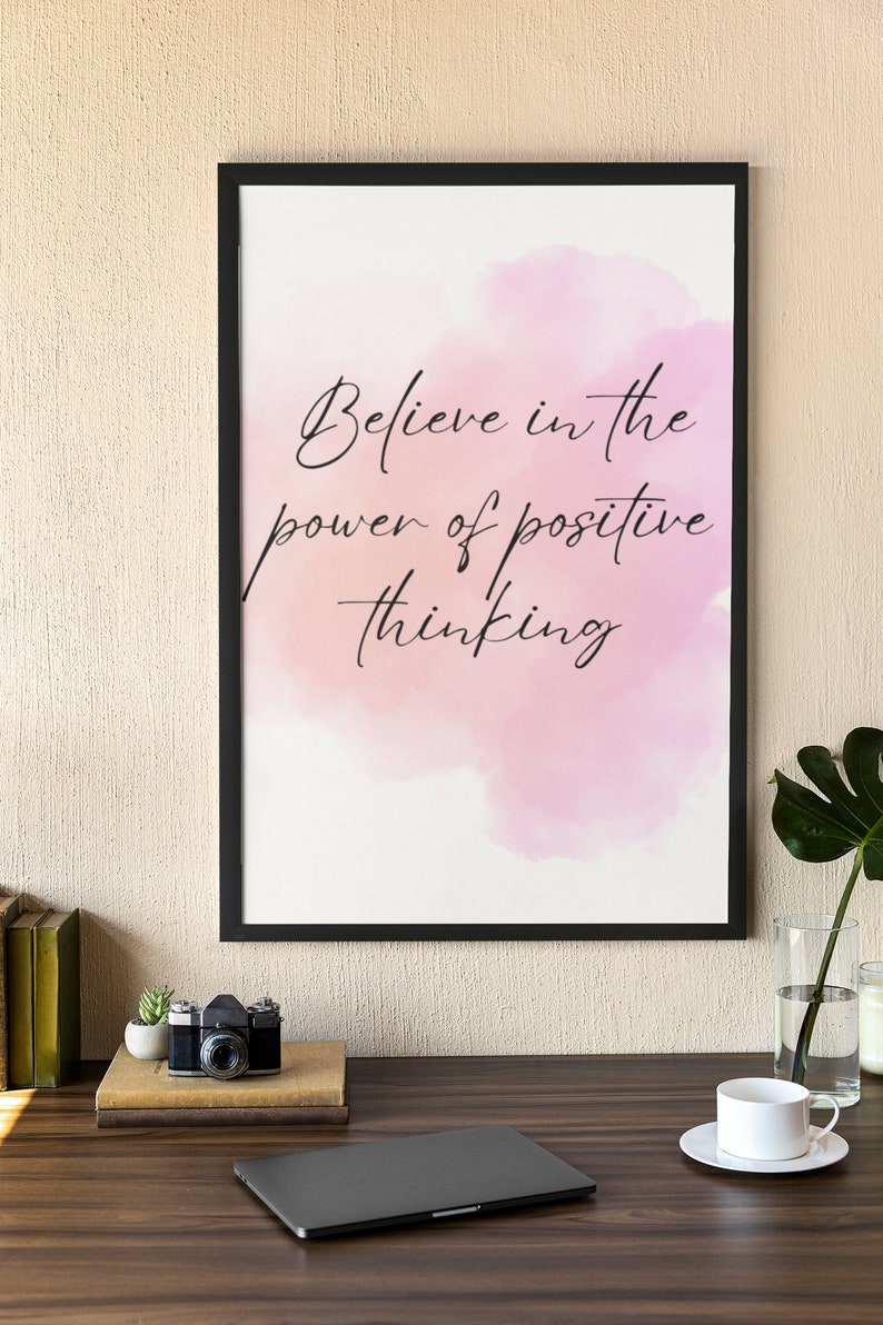 Peut inclure: Une impression encadr&eacute;e avec un fond aquarelle rose et le texte "Croyez au pouvoir de la pens&eacute;e positive". L'impression est sur un bureau avec un ordinateur portable, un appareil photo, une plante en pot et une tasse et une soucoupe.