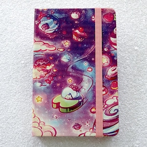 Peut inclure: Un carnet rose et violet avec un motif sur le thème de la galaxie représentant une variété de bonbons colorés et d'étoiles. Le carnet est doté d'une fermeture élastique rose.