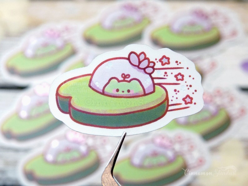 May include: A die-cut sticker featuring a cartoon avocado with a frog inside a pink spaceship. The avocado is green with a brown pit, and the spaceship has pink accents and stars. The sticker has a white border.