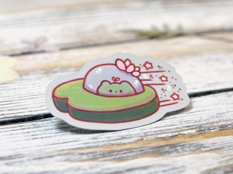 May include: A sticker featuring a cute green frog in a UFO-shaped vehicle. The UFO is green and gray with a red outline, and the frog is inside a clear dome. Pink stars and a flower accent the design.