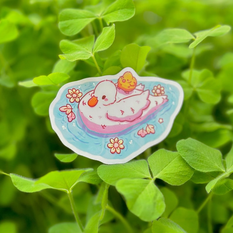 May include: A sticker featuring a white duck with a yellow chick on its back, swimming in a blue pond with pink and yellow flowers. The sticker is on a green background.