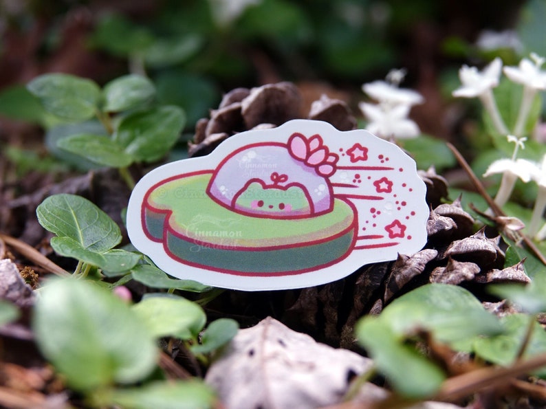 May include: A whimsical sticker featuring a green frog in a spaceship-like vehicle. The vehicle is green and brown, with a pink dome and pink stars trailing behind. The sticker has a white border and is set against a natural background.