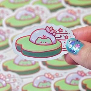 May include: A die-cut sticker featuring a cute cartoon frog in a UFO-shaped avocado. The UFO is light green with a red border, and the frog is light green with a pink blush. The sticker has a white background and is held by a hand.