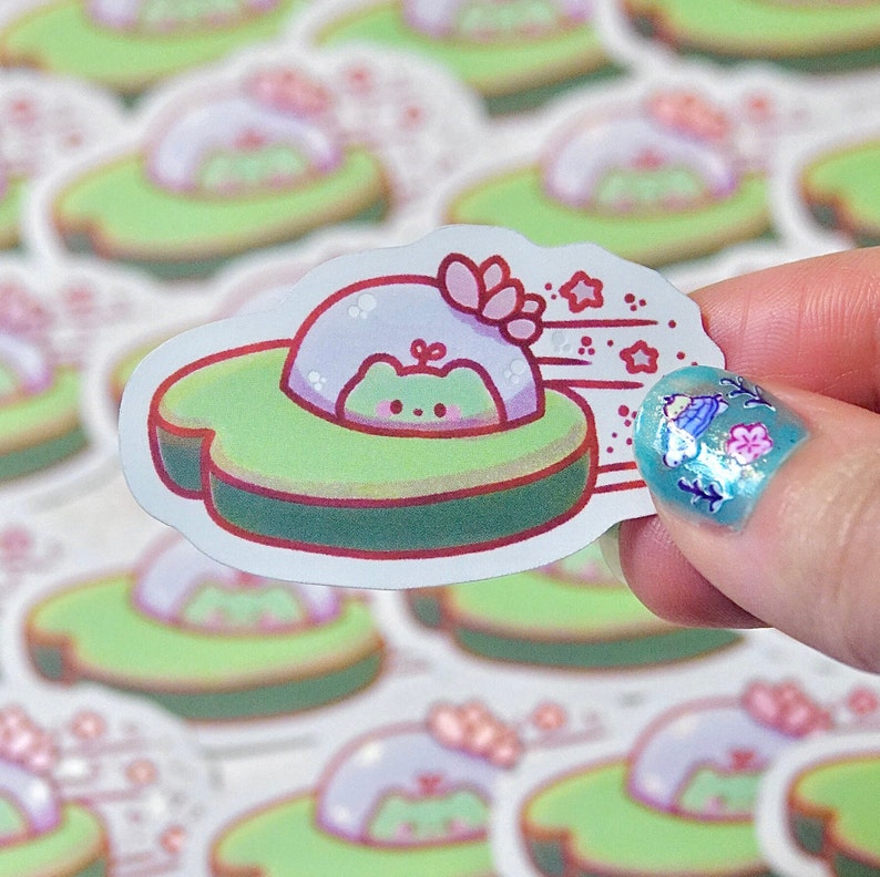May include: A die-cut sticker featuring a cute cartoon frog in a UFO-shaped avocado. The UFO is light green with a red border, and the frog is light green with a pink blush. The sticker has a white background and is held by a hand.