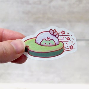 May include: A sticker featuring a cartoon frog in a UFO. The UFO is green and red, with a white and pink object on top. Red stars and lines indicate movement. The sticker has the word "cinnamon" on it.