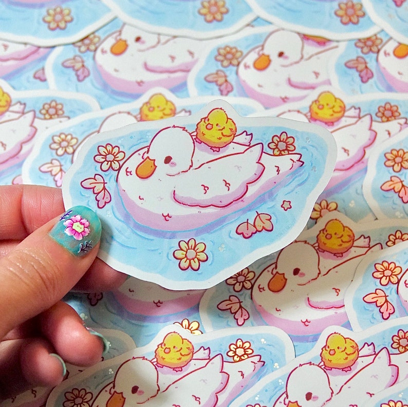 May include: A set of stickers featuring cute cartoon illustrations of swans with baby swans. The stickers are on a white background with blue and pink accents. The swans are surrounded by flowers and leaves.