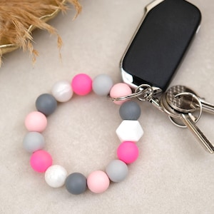 Könnte beinhalten: Ein Armband-Schlüsselanhänger in Kreisform. Das Armband hat eine Kombination aus rosa, grau, weiß und einer einzelnen weißen geometrischen Perle. Ein schwarzer Autoschlüsselanhänger und Schlüssel liegen neben dem Armband.