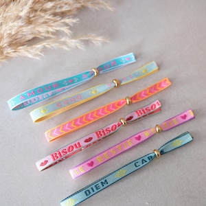 Könnte beinhalten: Eine Sammlung farbenfroher Freundschaftsarmbänder mit goldfarbenen Verschlüssen. Jedes Armband hat eine andere Farbe und Text, darunter "AMOUR", "GOOD", "Bisou", "BESTIE" und "CARPE DIEM". Die Armbänder sind aus gewebtem Stoff.