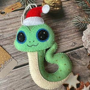 May include: Green felt snake Christmas ornament with a white felt Santa hat. The snake has big black eyes and a stitched smile. The ornament has a red heart with the text "Handmade with love".