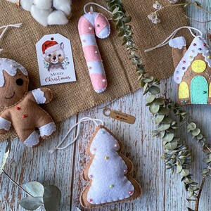 May include: Four felt Christmas decorations: a gingerbread person, a candy cane, a gingerbread house, and a Christmas tree. Each decoration is decorated with white icing and colourful sprinkles. The decorations are hanging from a hessian background.